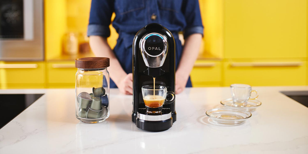 Woman stands behind a pod machine while it pours an artisan coffee into a glass espresso cup