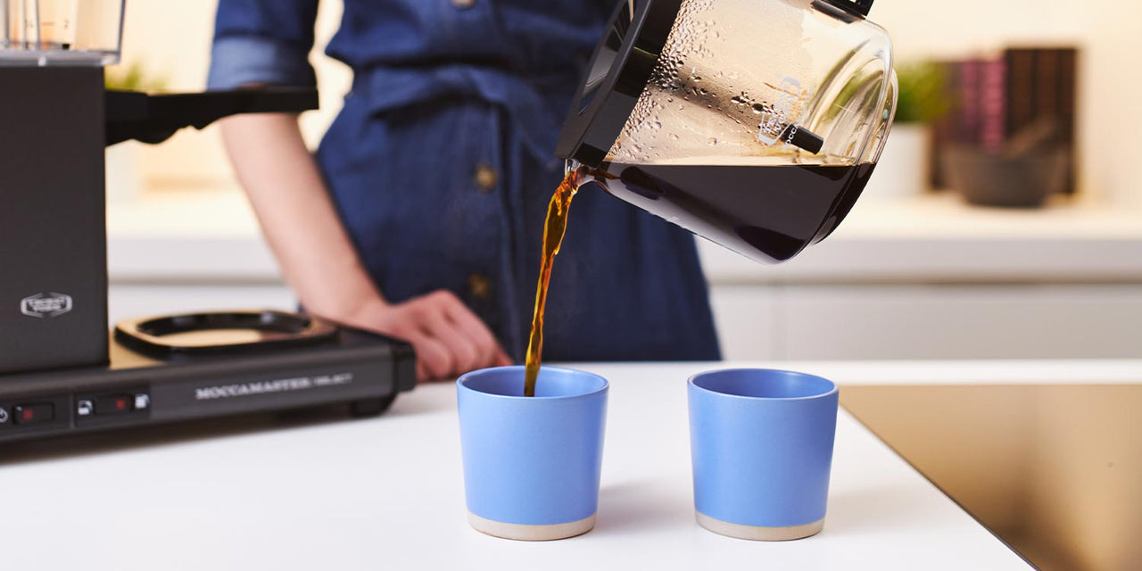 Woman pours artisan coffee from moccamaster machine into two blue ceramic cups