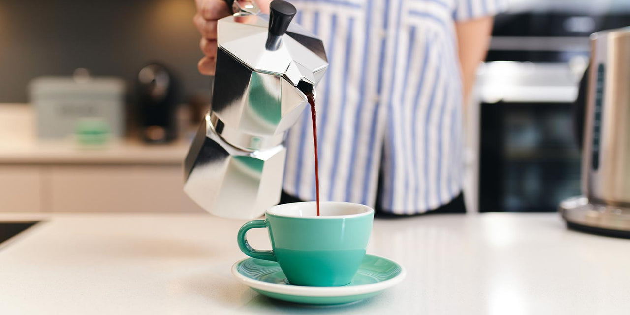 Woman pours artisan coffee out of a silver moka pot into a green cup with saucer