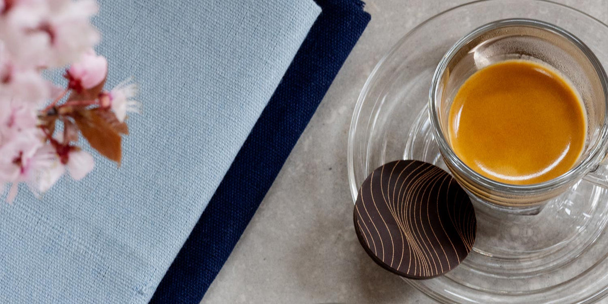 A glass espresso cup and saucer holds an Artisan Coffee co coffee with a perfect crema. On the saucer is a chocolate disc. To the left is a dark blue napkin with a light blue napkin on top and in the top left corner pink flowers are slightly out of focus