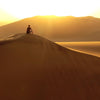 A man sits with his back to the camera on a large sand dune watching the sun set over another sand dune in the distance