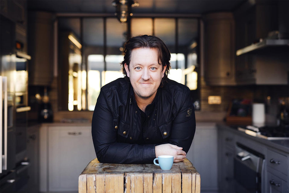 Ashley Palmer-Watts, cofounder of Artisan Coffee Co leans forward on a wooden surface mid frame. In the background is a kitchen with an oven, fridge and large window. He has wavy dark hair, is smiling and has a blue espresso mug invfront of him