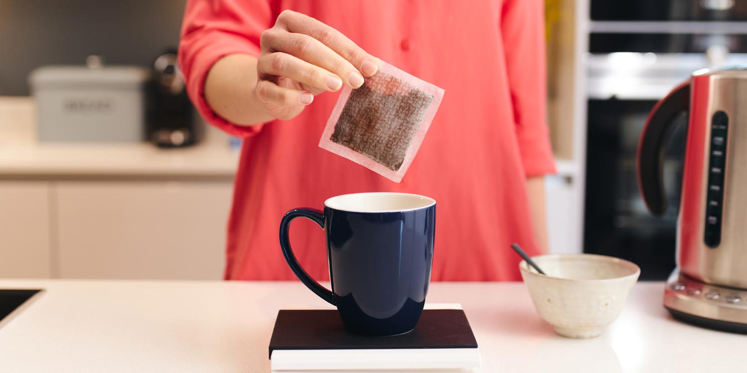 Woman in a red shirt putting an artisan coffee bag into a black mug which is on top of a set of digital scales. A pot of sugar and kettle are beside the mug