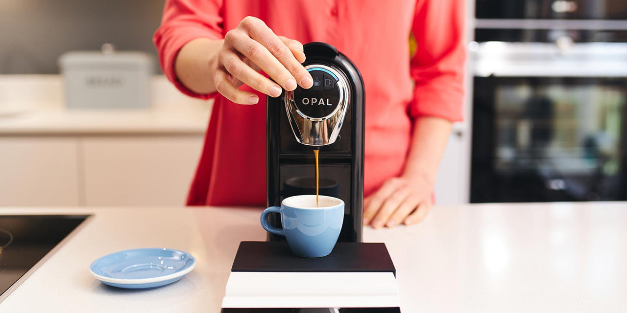 A woman in a red shirt holds the button down on her coffee pod machine while artisan coffee pours into a blue ceramic cup from the same machine. The cup sits on a set of digital scales and the saucer is to the left of the machine.