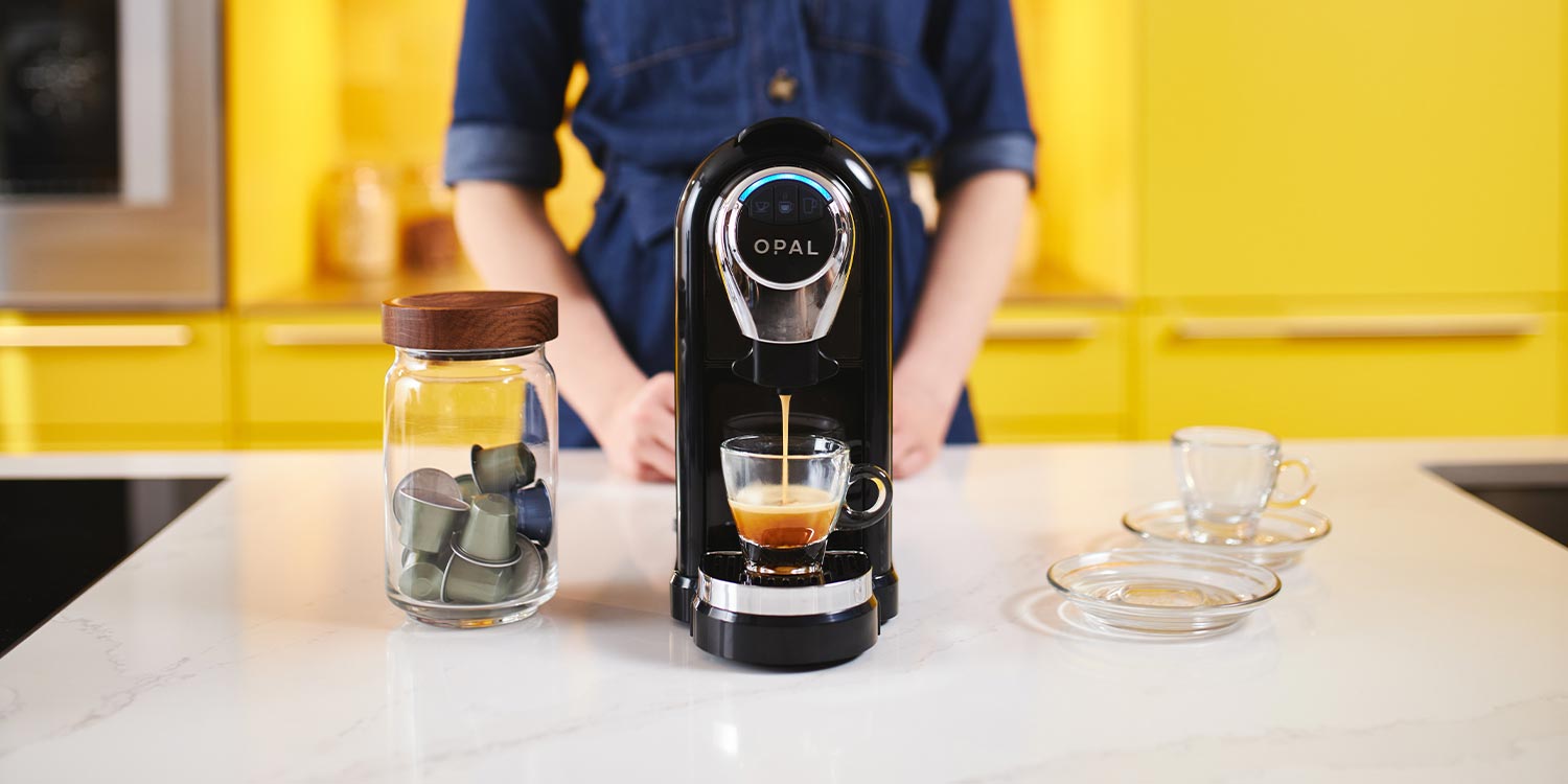 Woman stands behind a pod machine while it pours an artisan coffee into a glass espresso cup 