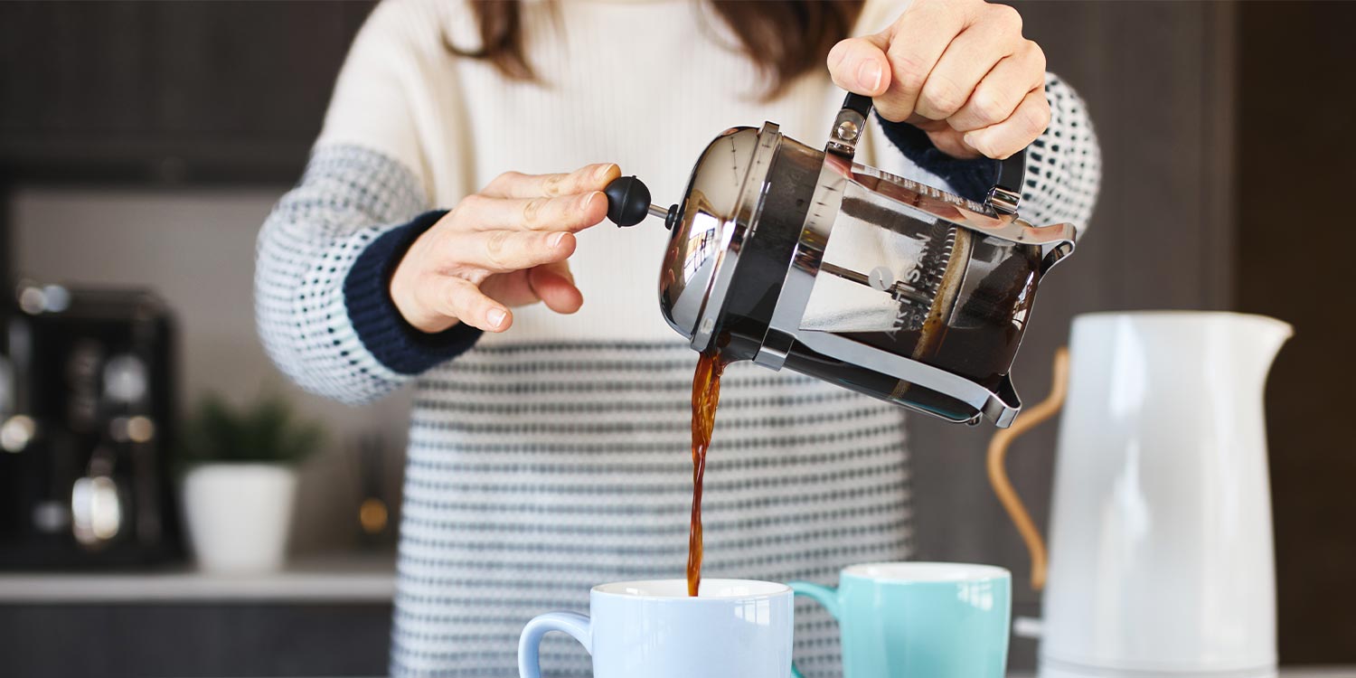 Woman pours artisan coffee from a cafetière into blue mug with jug of water next to her 