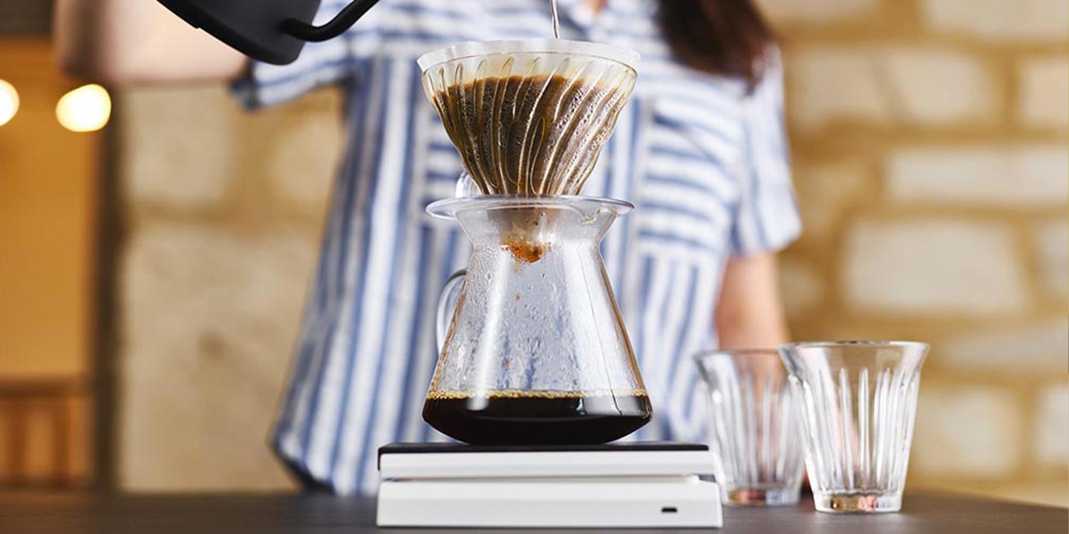 woman is striped top pours water into a V60 pour over vessel through a paper filter on top of scales