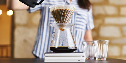 woman is striped top pours water into a V60 pour over vessel through a paper filter on top of scales