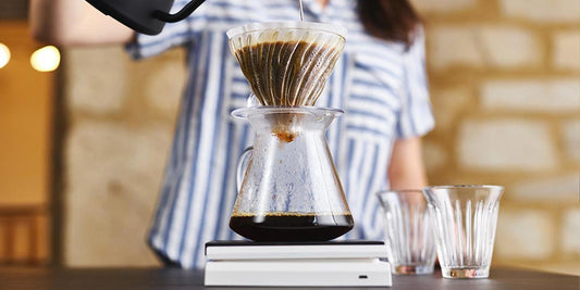 woman is striped top pours water into a V60 pour over vessel through a paper filter on top of scales