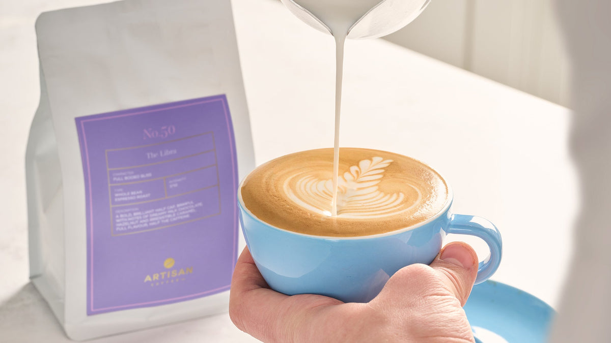 Latte art being poured into baby blue coloured cup in front of a bag of Libra Whole Bean coffee sitting on counter top