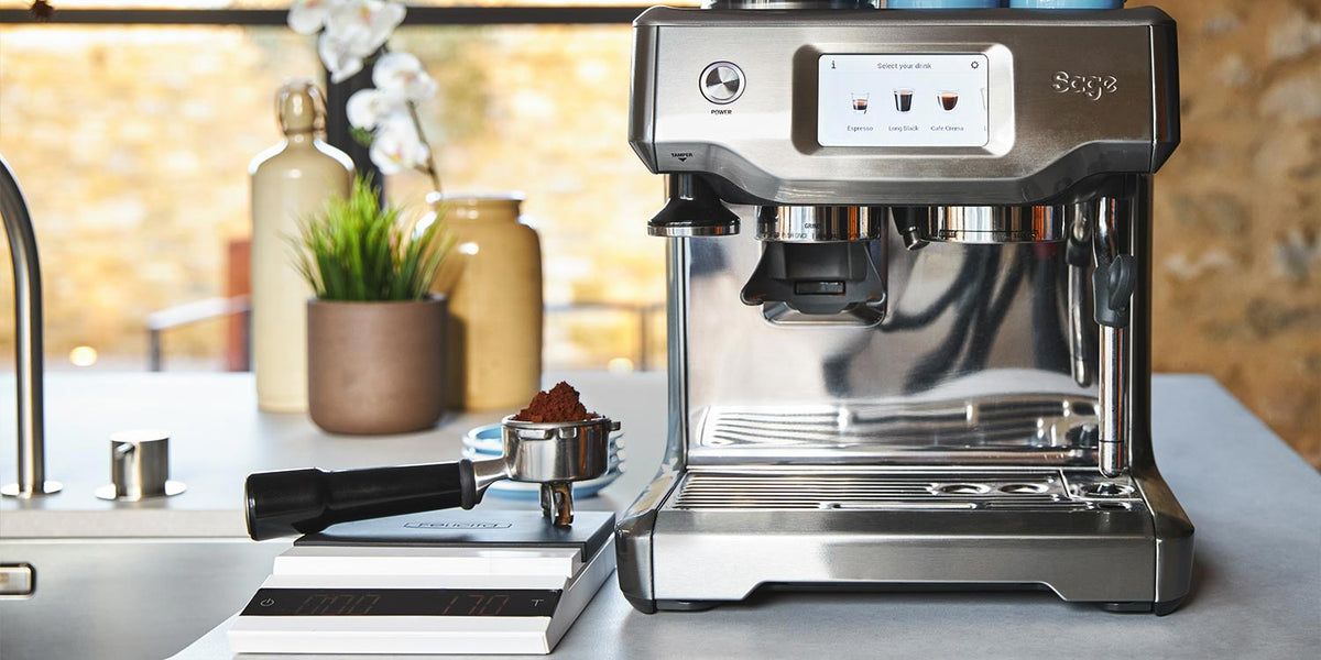 An espresso machine pod filled with Artisan Coffee Co coffee rests on a set of digital scales infront of the machine. There are ceramic vases in the background with flowers and grass like plants in them