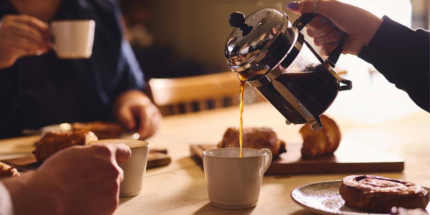 Artisan coffee being poured from a cafetiere into a mug on a table as friends sit around a table to chat and drink coffee.