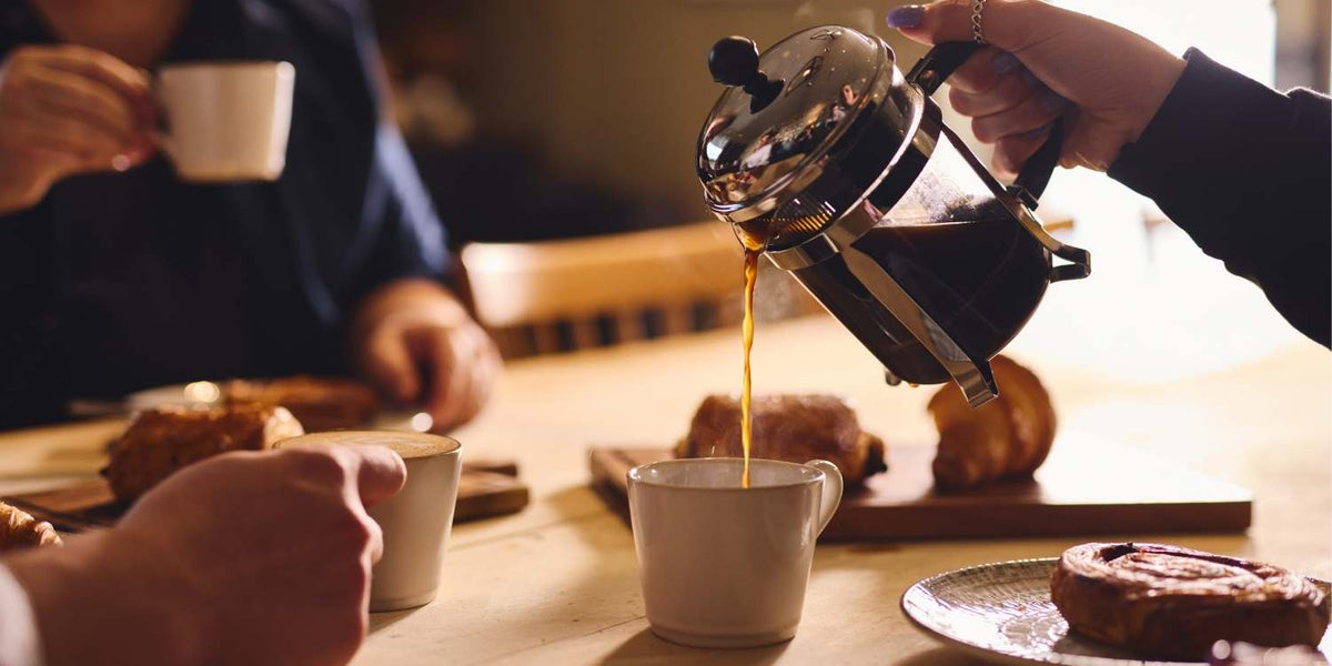 Artisan coffee being poured from a cafetiere into a mug on a table as friends sit around a table to chat and drink coffee.
