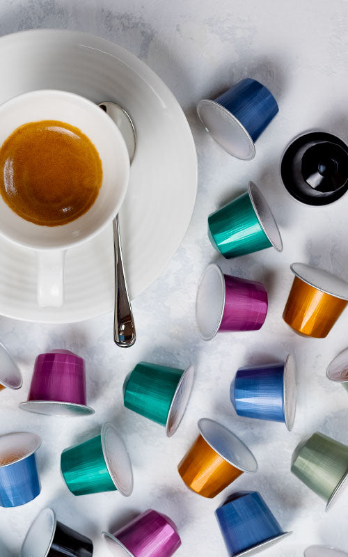 An assortment of various coloured pod capsules next to a mug of coffee on a white table